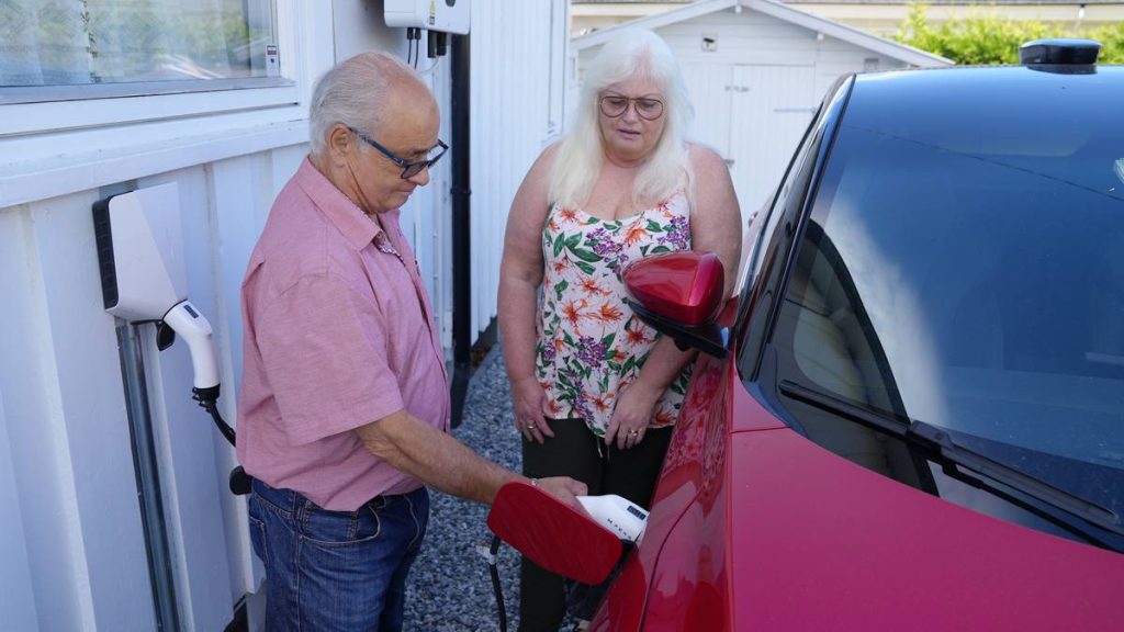 En mann og en kvinne står ved en rød elbil og mannen setter strømledningen i bilen.